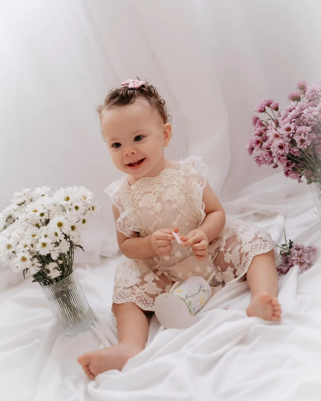 Bebê de vestido de renda sentada entre flores brancas e lilás