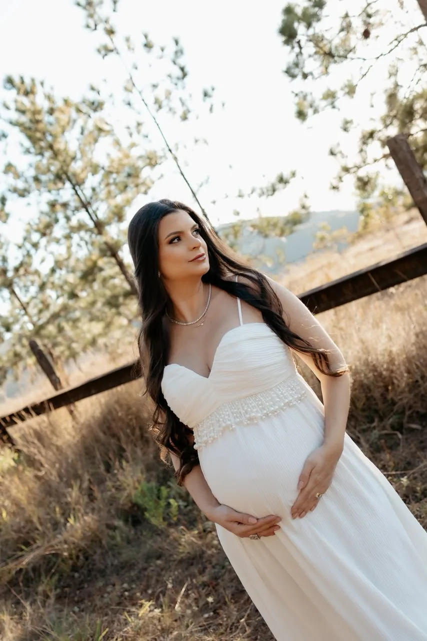Gestante de vestido branco com as mãos na barriga, num campo ao fim da tarde