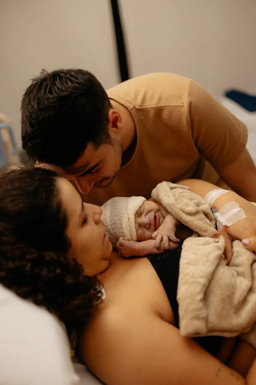 Mãe recebe o bebê no colo logo após o nascimento, com o pai ao lado
