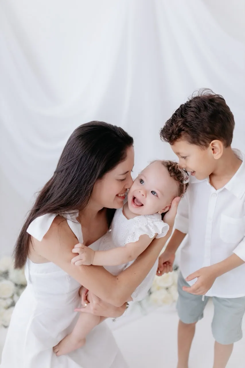 Mãe beija a bebê no colo enquanto o irmão mais velho sorri