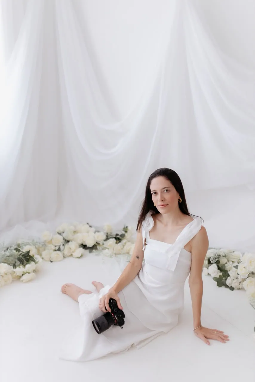 Isabella Miranda sentada no estúdio, de vestido branco, com a câmera entre rosas brancas